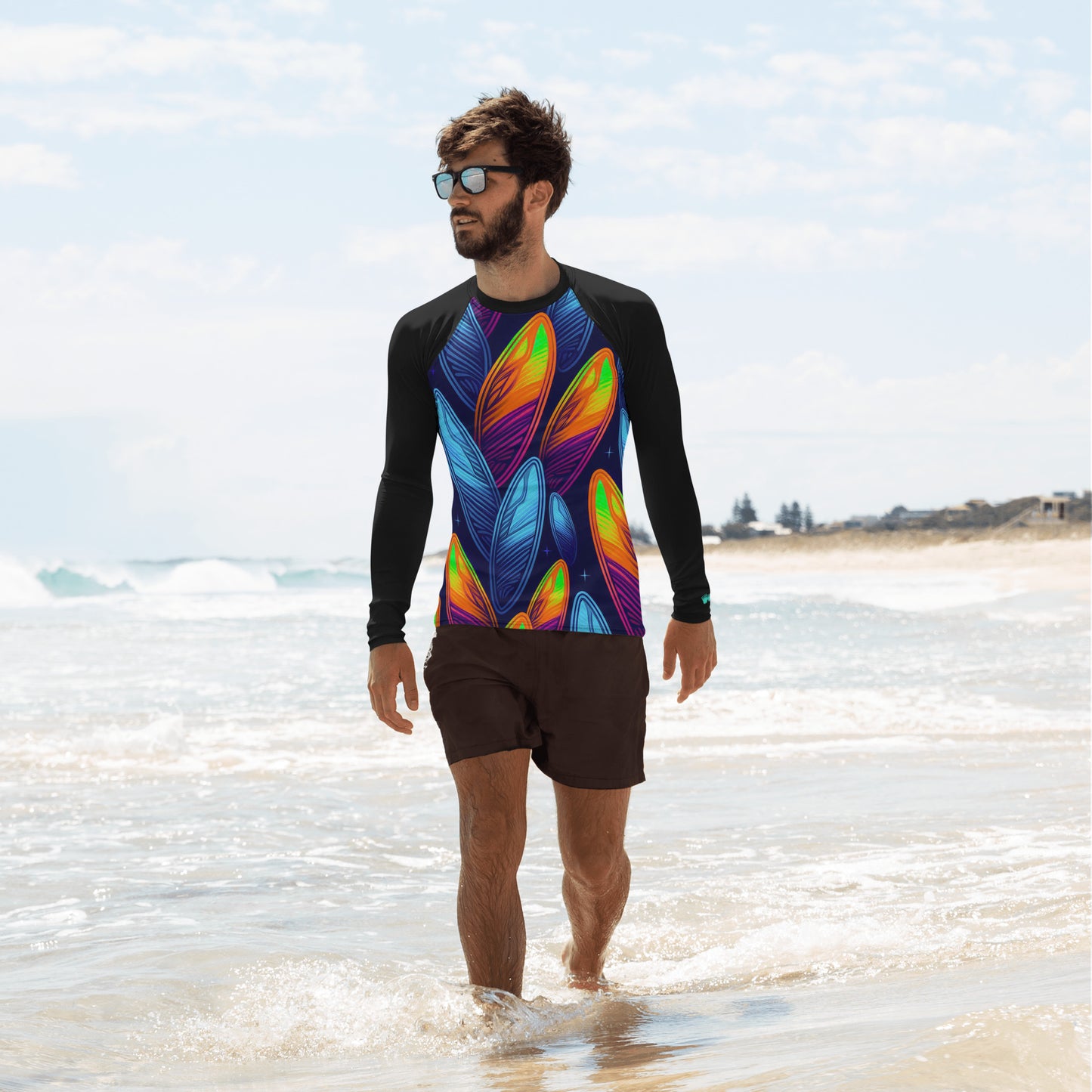 Stylish man in colorful rash guard walking on the beach, showcasing vibrant surfboard pattern and sun protection.